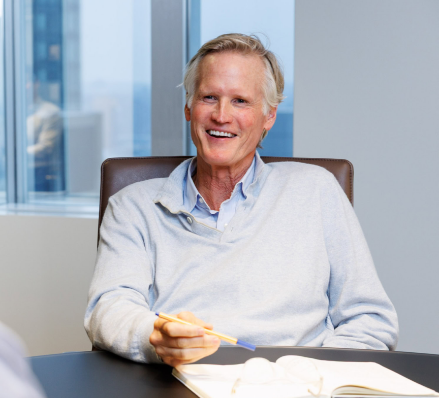 Charlie Gifford, an older man with gray hair, sits at a table in a modern office, smiling as he holds a pen over his notebook. Glasses and a phone rest on the table before him, with large windows in the background.
