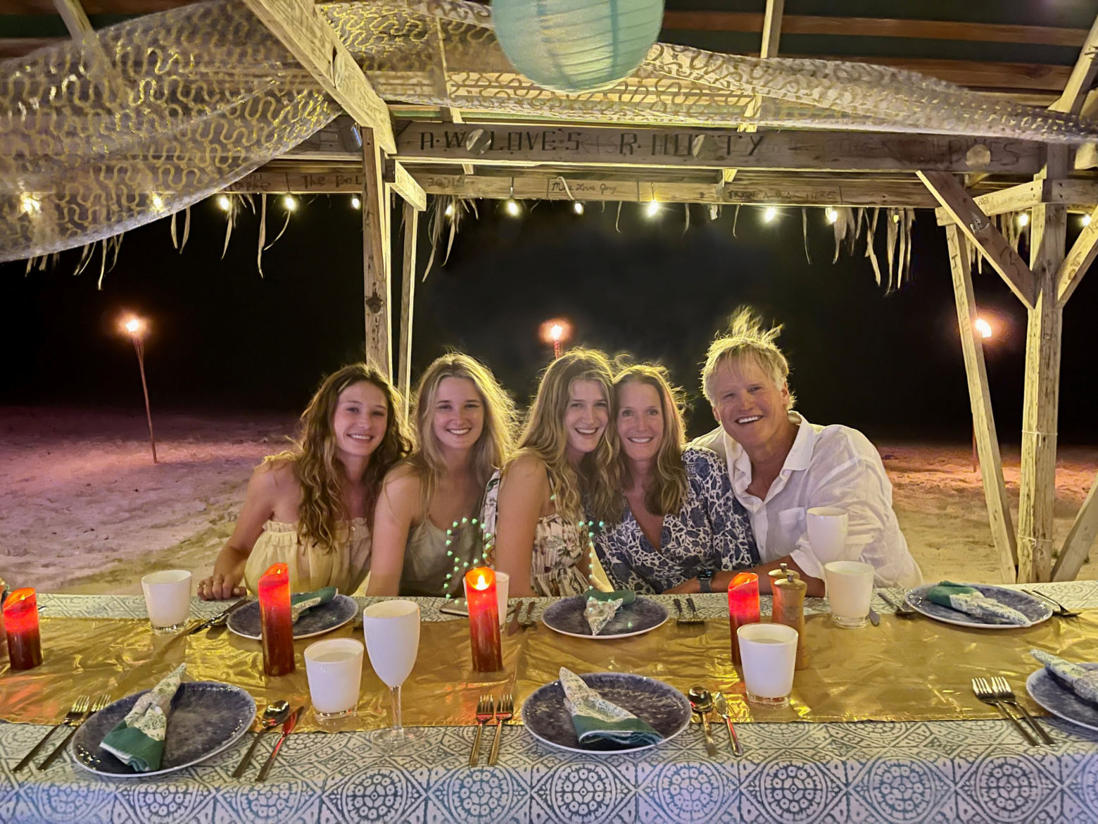 Five people, including Charlie Gifford, sit closely together, smiling at a beachside table set for dinner under string lights and a wooden canopy at night. The table is decorated with plates, napkins, candles, and cups.