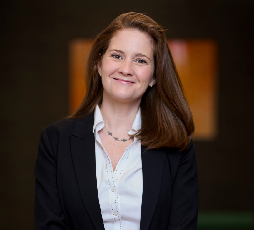 Melissa Barry, a woman with long brown hair, wearing a black blazer over a white blouse, stands smiling in front of a blurred dark background with warm-colored rectangular shapes.