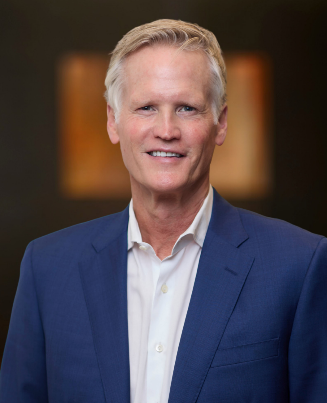 Charlie Gifford, a man with short blond hair wearing a blue suit jacket and white dress shirt, smiles at the camera against a blurred, dark background.