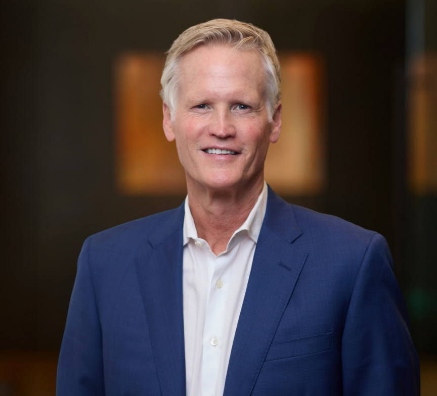 Charlie Gifford, a man with short blond hair wearing a blue suit jacket and white dress shirt, smiles at the camera against a blurred, dark background.
