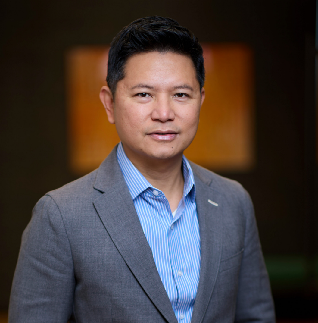 Tristan Velez stands confidently in a gray suit jacket and blue striped shirt, looking at the camera with a slight smile. The background is softly blurred with warm colors.