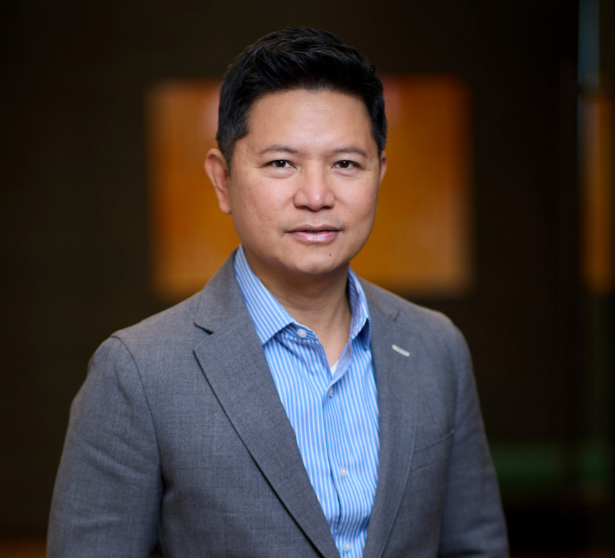 Tristan Velez stands confidently in a gray suit jacket and blue striped shirt, looking at the camera with a slight smile. The background is softly blurred with warm colors.