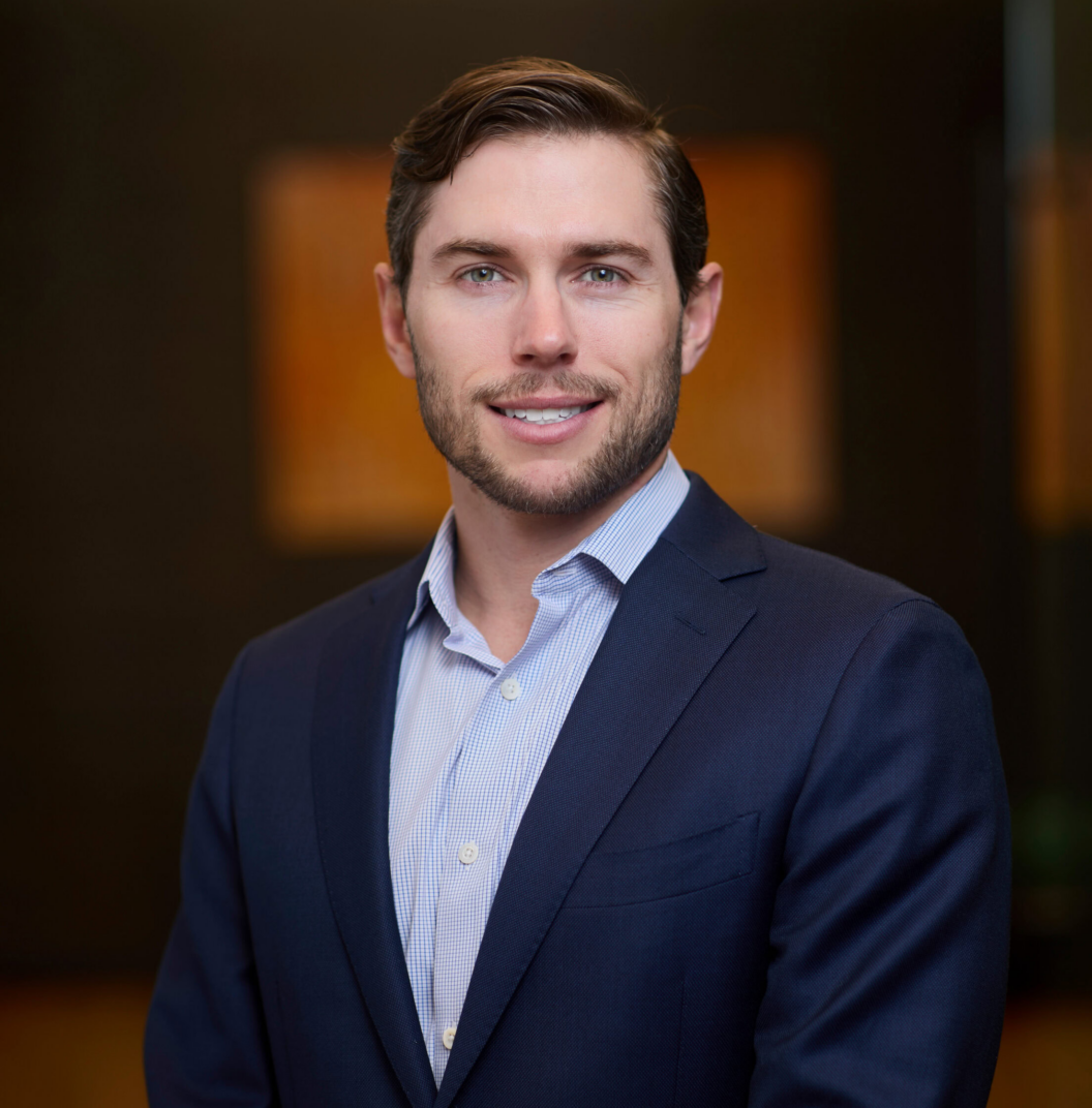 A man with short brown hair and a trimmed beard, wearing a navy suit and light blue shirt, stands indoors in front of a blurred dark background. He is smiling and looking at the camera.