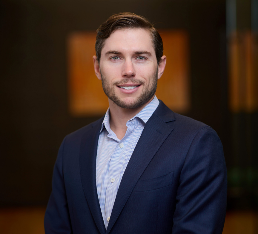 A man with short brown hair and a trimmed beard, wearing a navy suit and light blue shirt, stands indoors in front of a blurred dark background. He is smiling and looking at the camera.