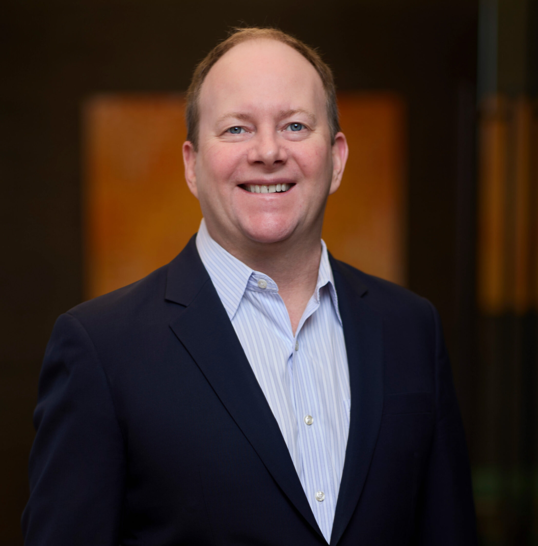 Judson Samuels, in a navy blue suit jacket and light blue dress shirt, stands smiling against a blurred dark background with warm tones.