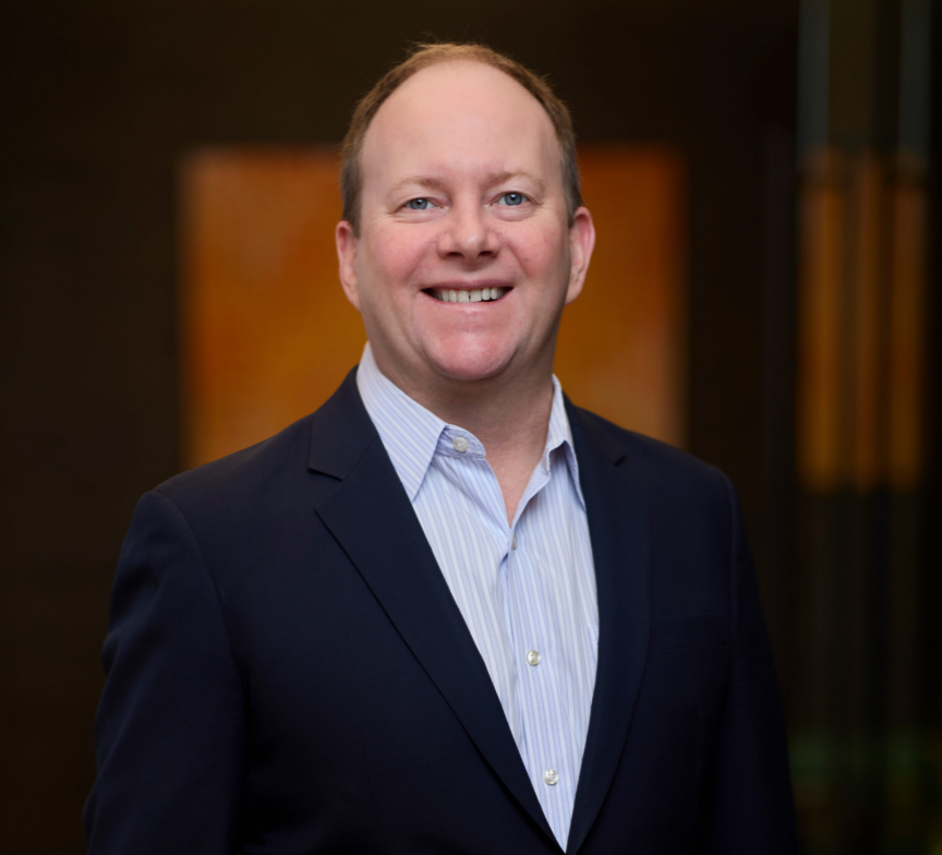Judson Samuels, in a navy blue suit jacket and light blue dress shirt, stands smiling against a blurred dark background with warm tones.