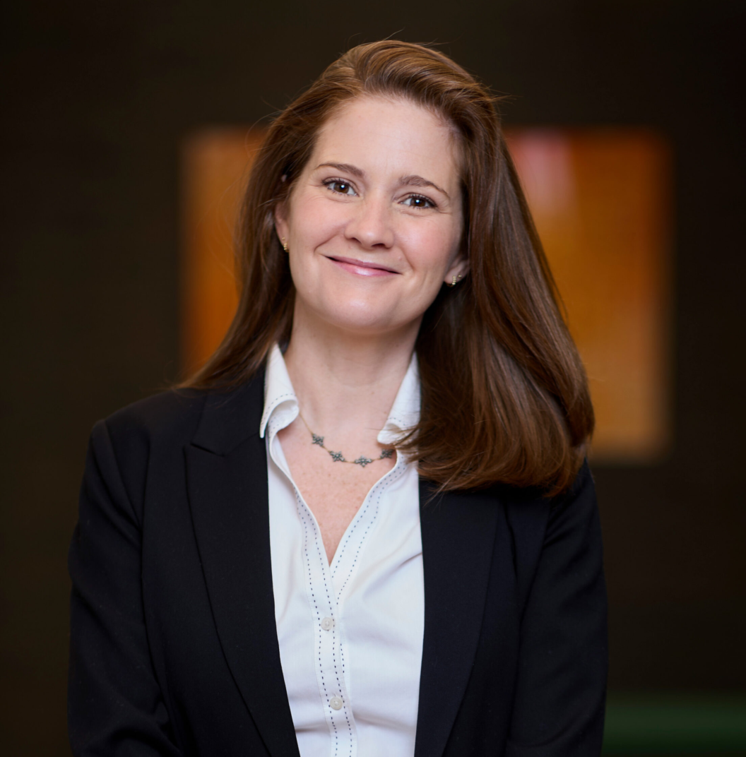 Melissa Barry, a woman with long brown hair, wearing a black blazer over a white blouse, stands smiling in front of a blurred dark background with warm-colored rectangular shapes.