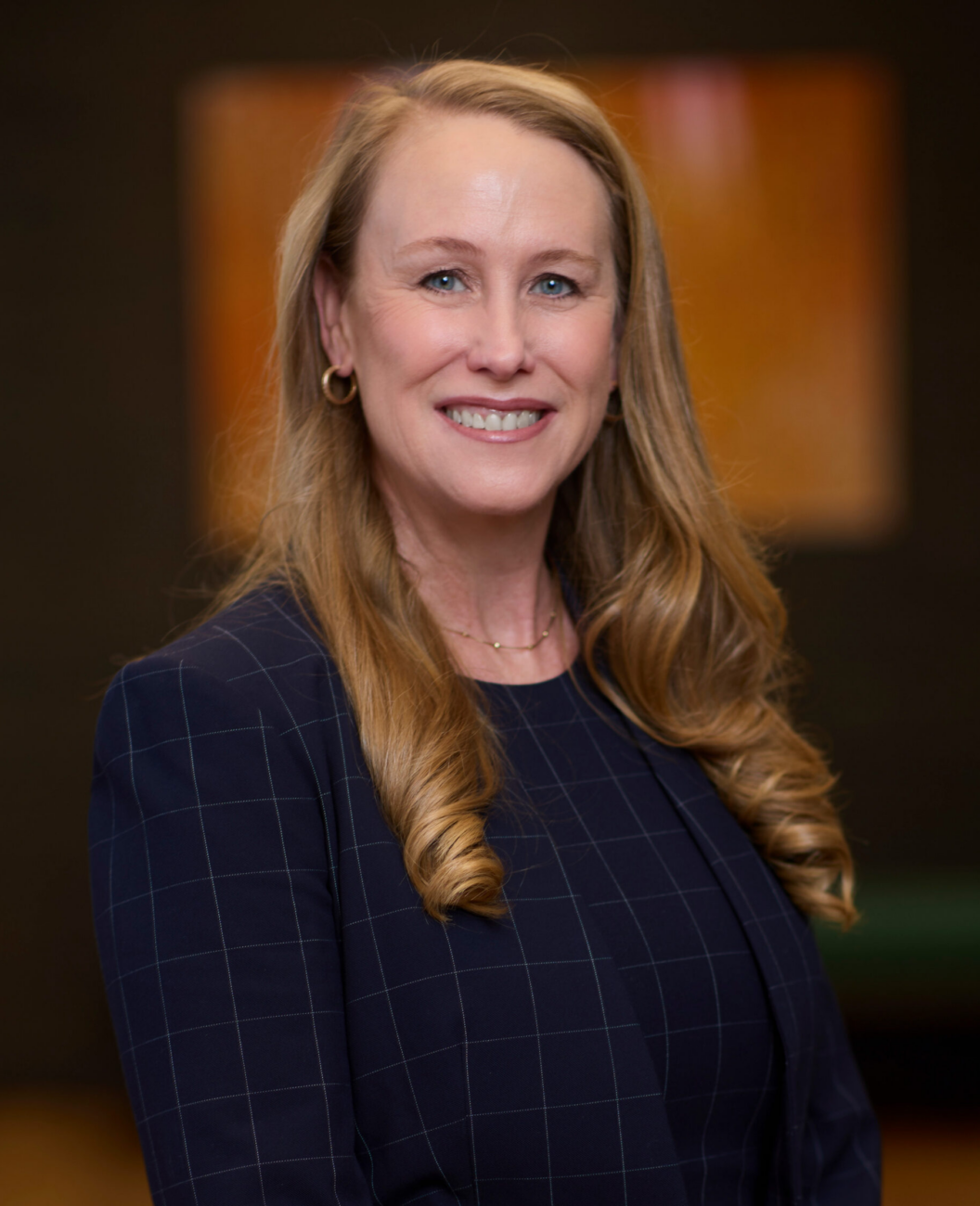 Nickie Norris, a woman with long blonde hair, wearing a navy blue checked blazer and a dark top, smiles while standing indoors with a softly blurred background.