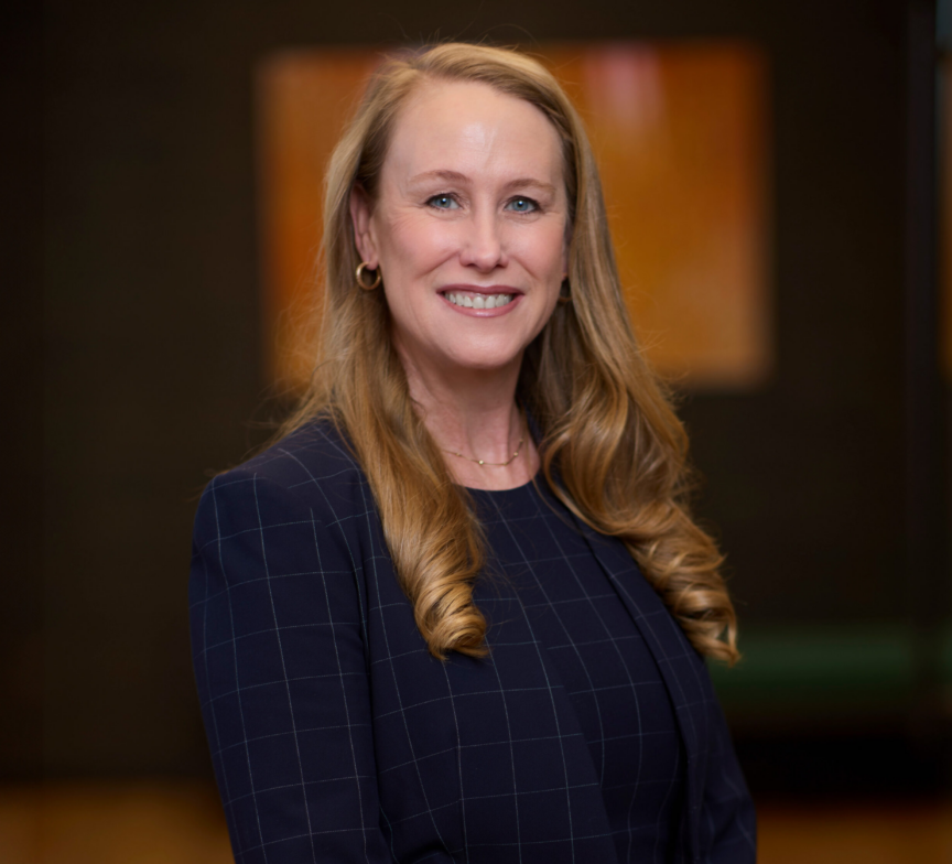 Nickie Norris, a woman with long blonde hair, wearing a navy blue checked blazer and a dark top, smiles while standing indoors with a softly blurred background.