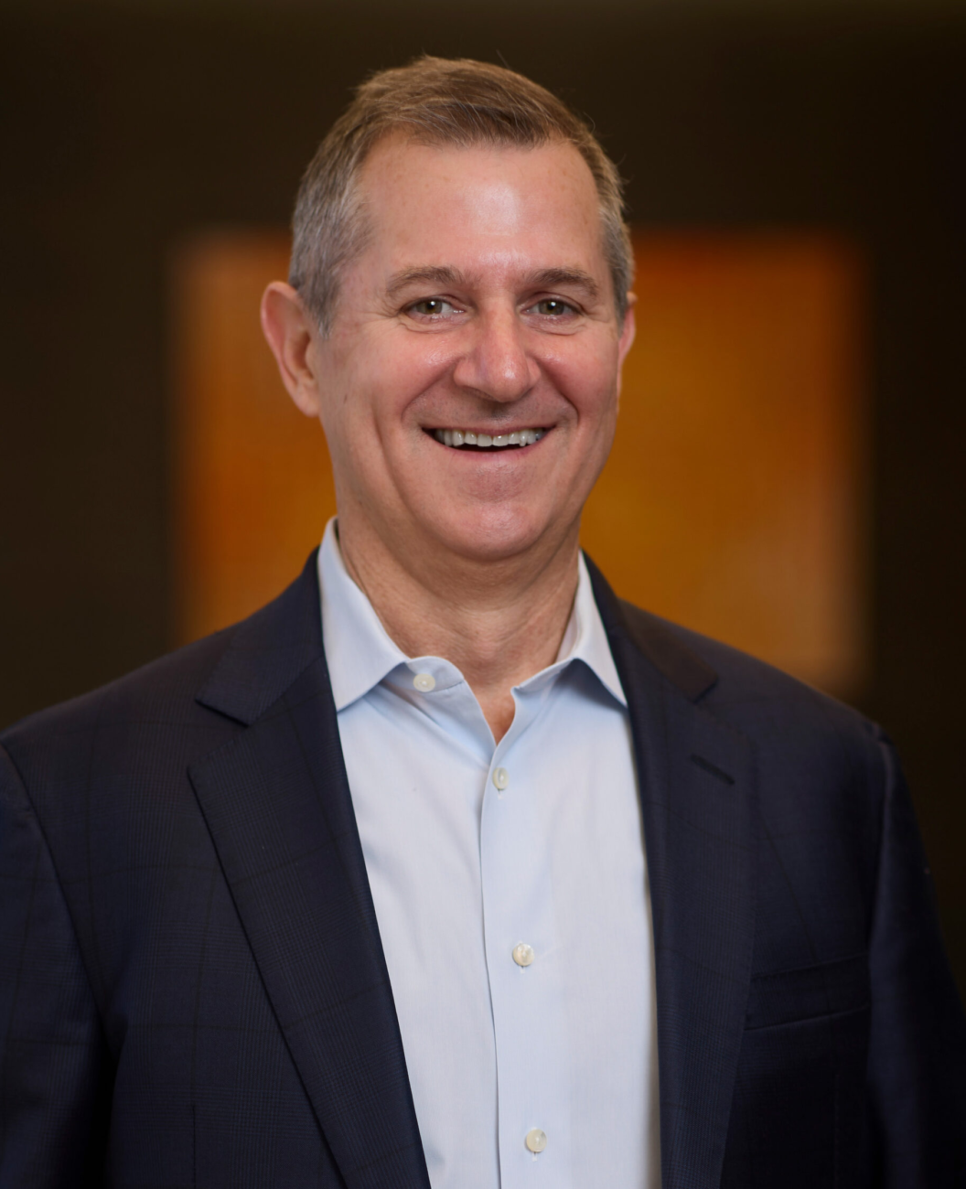 A smiling man wearing a dark suit jacket and light blue dress shirt stands in front of a softly blurred background with warm tones.