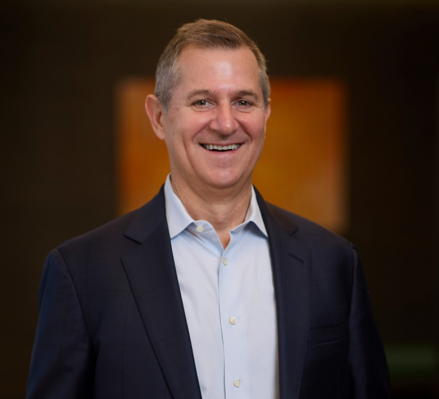 A smiling man wearing a dark suit jacket and light blue dress shirt stands in front of a softly blurred background with warm tones.