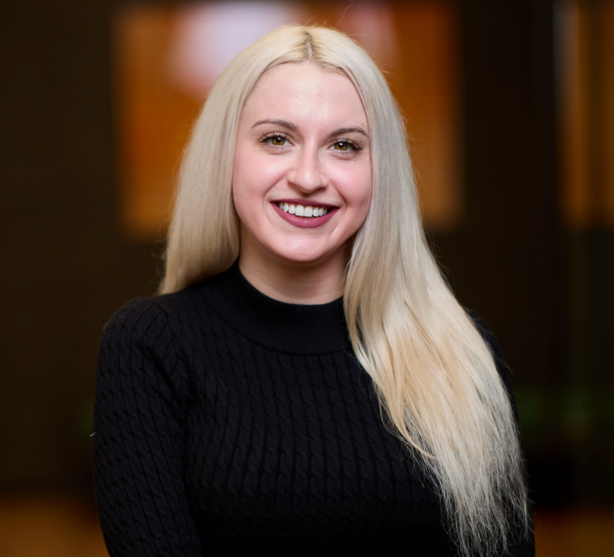A smiling woman with long, straight blonde hair wears a black sweater and stands indoors with a blurred, warm-toned background. This is Sarah Kalousdian.
