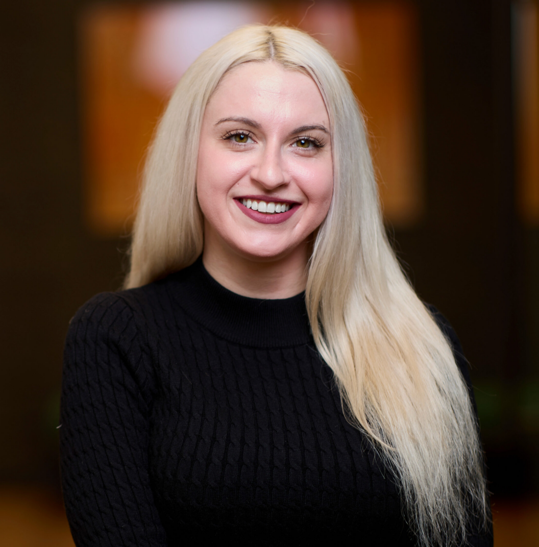 A smiling woman with long, straight blonde hair wears a black sweater and stands indoors with a blurred, warm-toned background. This is Sarah Kalousdian.