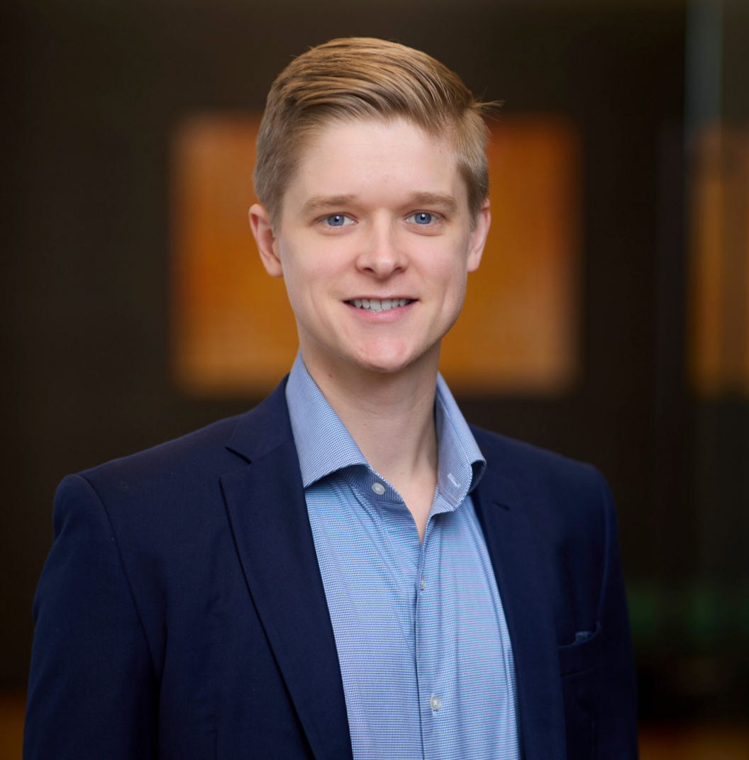 Bret Kuchenbecker, a young man in a blue suit jacket and light blue shirt, smiles at the camera. The background is softly blurred, showing a modern indoor setting with warm lighting and abstract wall art.