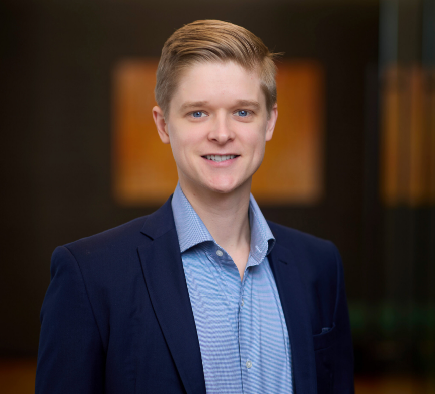 Bret Kuchenbecker, a young man in a blue suit jacket and light blue shirt, smiles at the camera. The background is softly blurred, showing a modern indoor setting with warm lighting and abstract wall art.