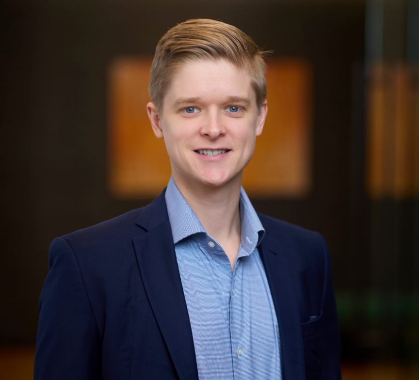 Bret Kuchenbecker, a young man in a blue suit jacket and light blue shirt, smiles at the camera. The background is softly blurred, showing a modern indoor setting with warm lighting and abstract wall art.