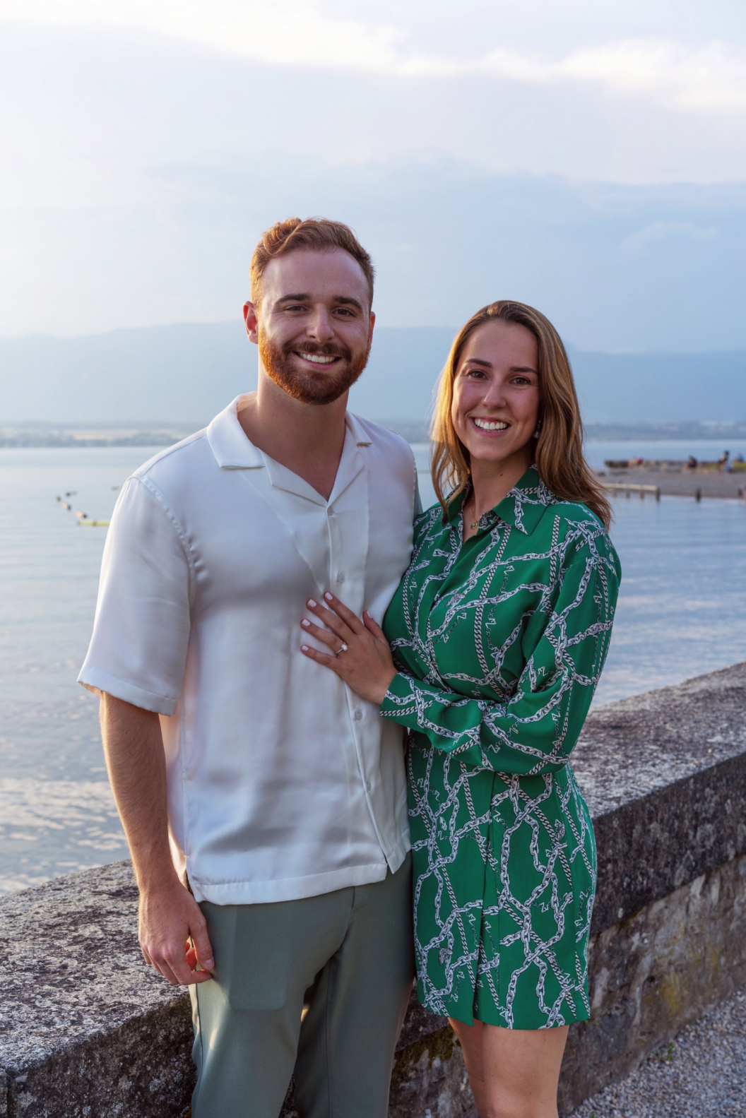 A smiling couple, including William Nemeth, poses together by a stone wall with a calm body of water and mountains in the background. The man wears a white shirt and green pants; the woman wears a green patterned dress.