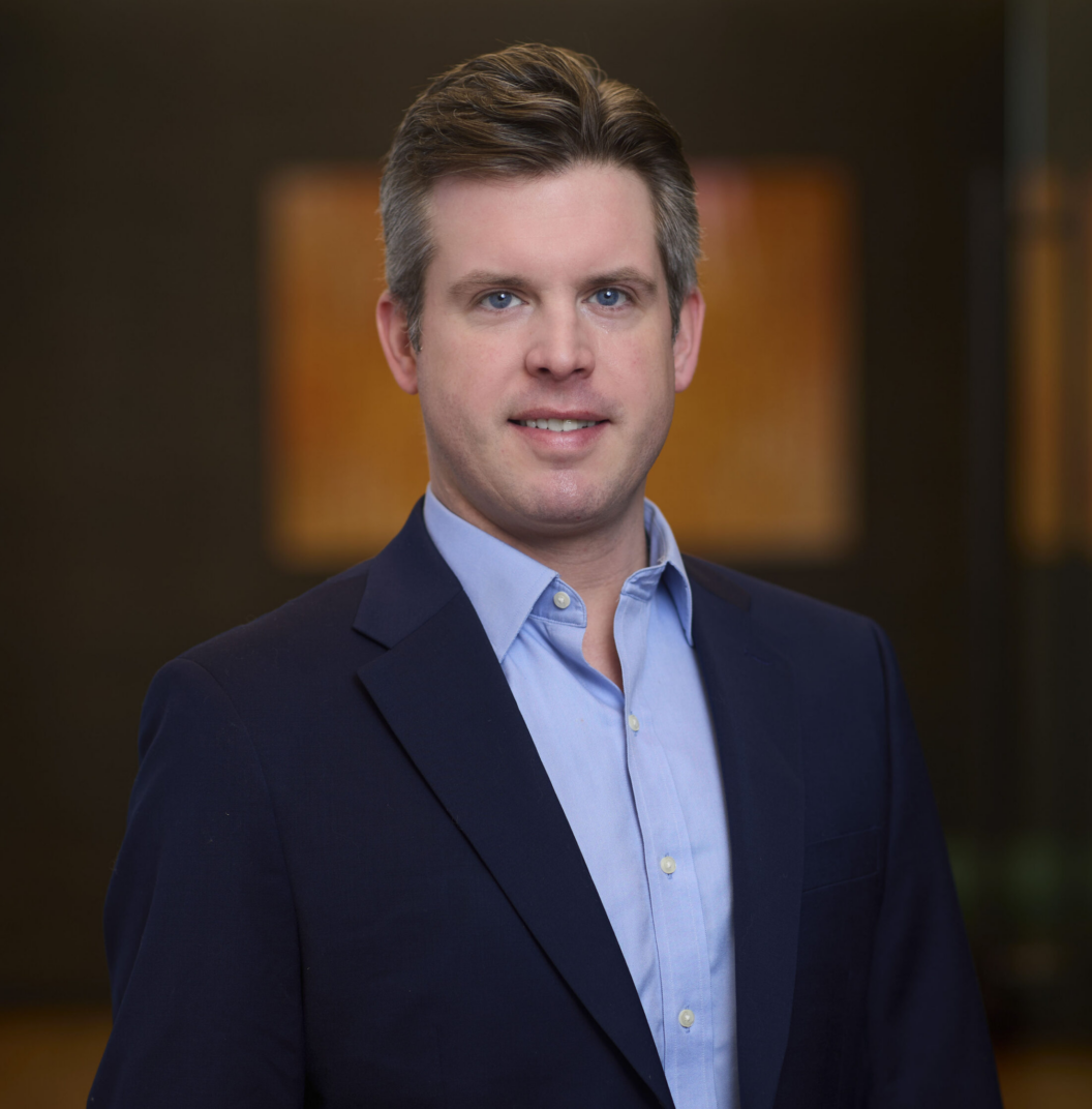 Scott MacPhee, a man with short brown hair, wearing a dark blue suit jacket and light blue dress shirt, stands indoors in front of a blurred, modern background with warm-toned artwork.
