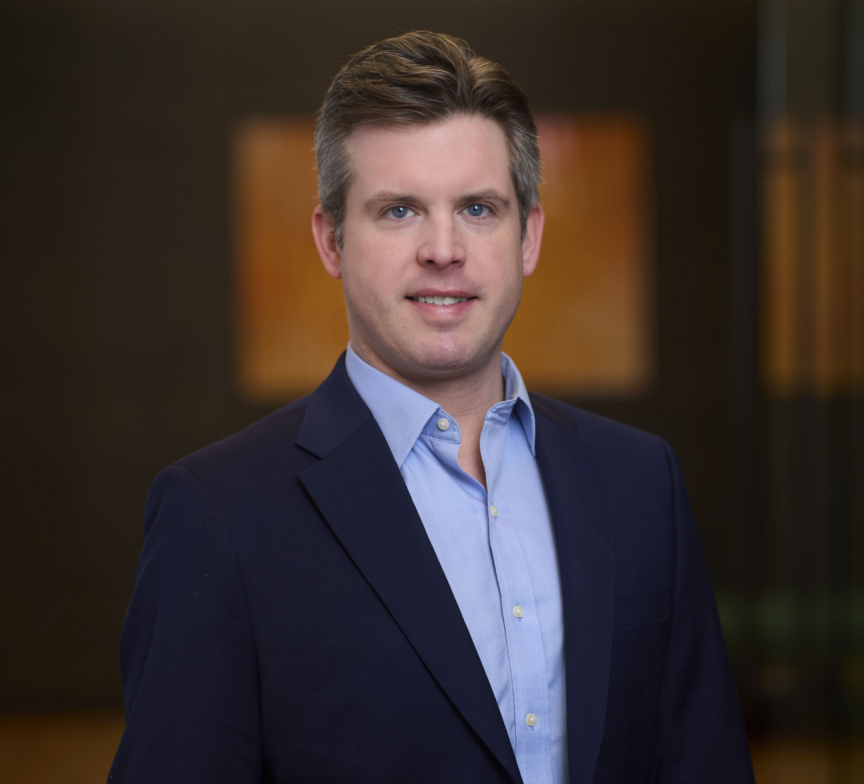 Scott MacPhee, a man with short brown hair, wearing a dark blue suit jacket and light blue dress shirt, stands indoors in front of a blurred, modern background with warm-toned artwork.