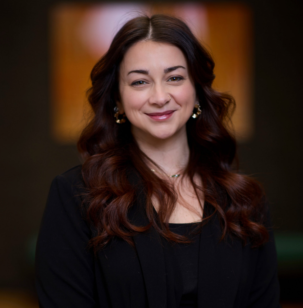 Tina O'Brien, with long, wavy dark hair and a warm smile, poses indoors in a black blazer and gold earrings. She stands against a softly blurred background with warm lighting.