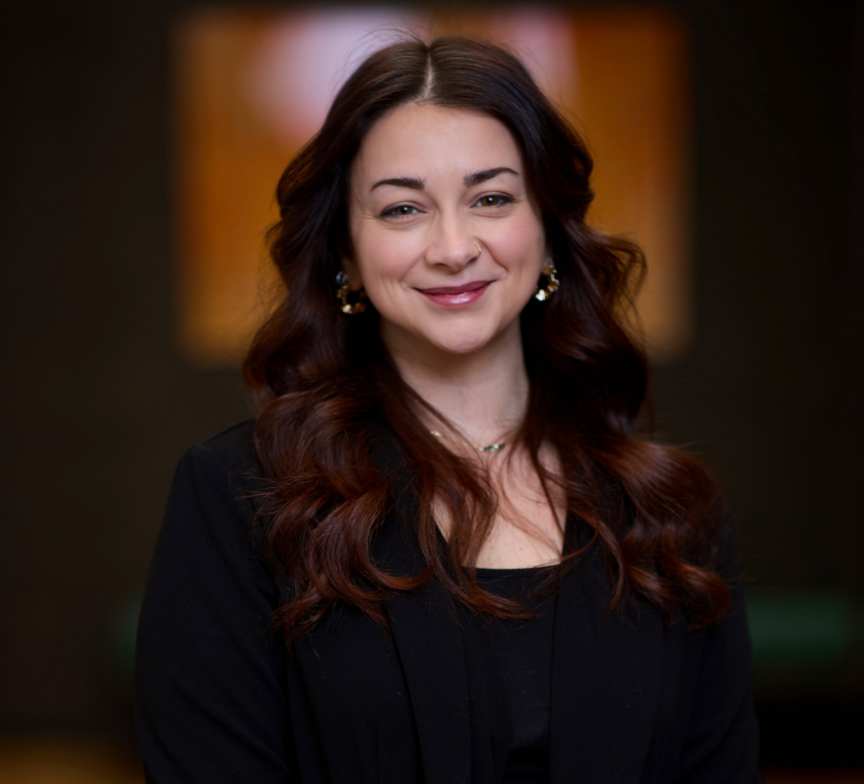Tina O'Brien, with long, wavy dark hair and a warm smile, poses indoors in a black blazer and gold earrings. She stands against a softly blurred background with warm lighting.