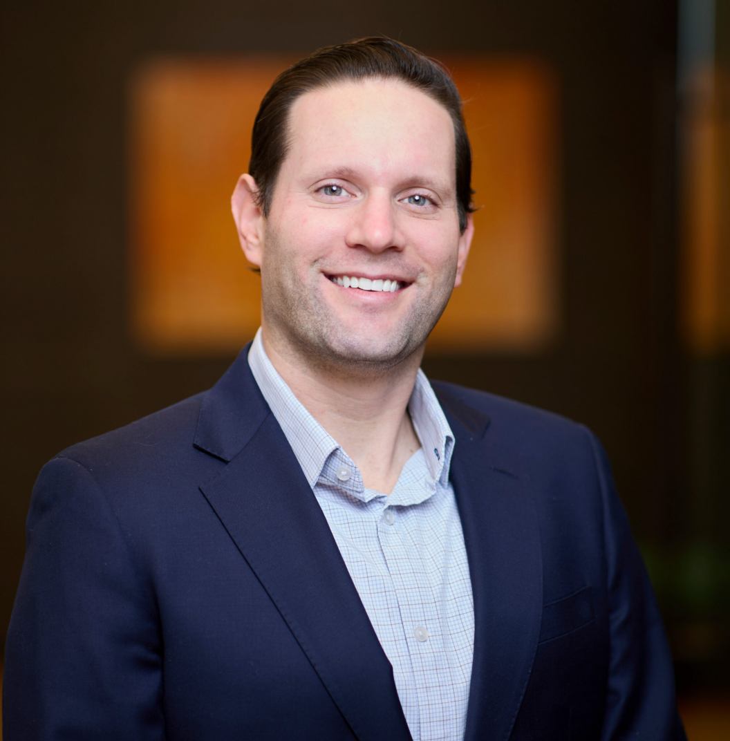 A man with light skin, dark hair, and a blue suit jacket over a light blue shirt smiles at the camera, standing indoors against a blurred dark background.