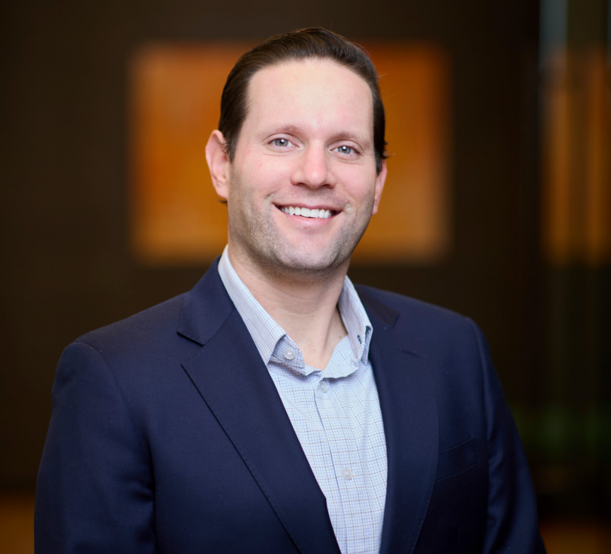 A man with light skin, dark hair, and a blue suit jacket over a light blue shirt smiles at the camera, standing indoors against a blurred dark background.