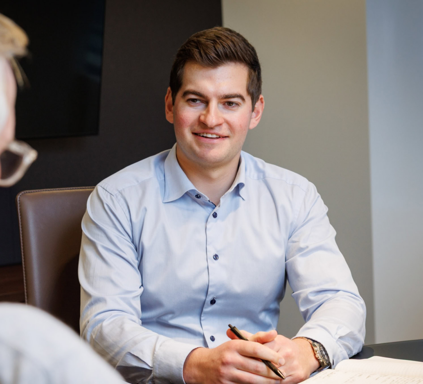 A man in a light blue dress shirt sits at a desk, smiling and holding a pen, with an open notebook in front of him. Another person is seated across from him, partially visible.