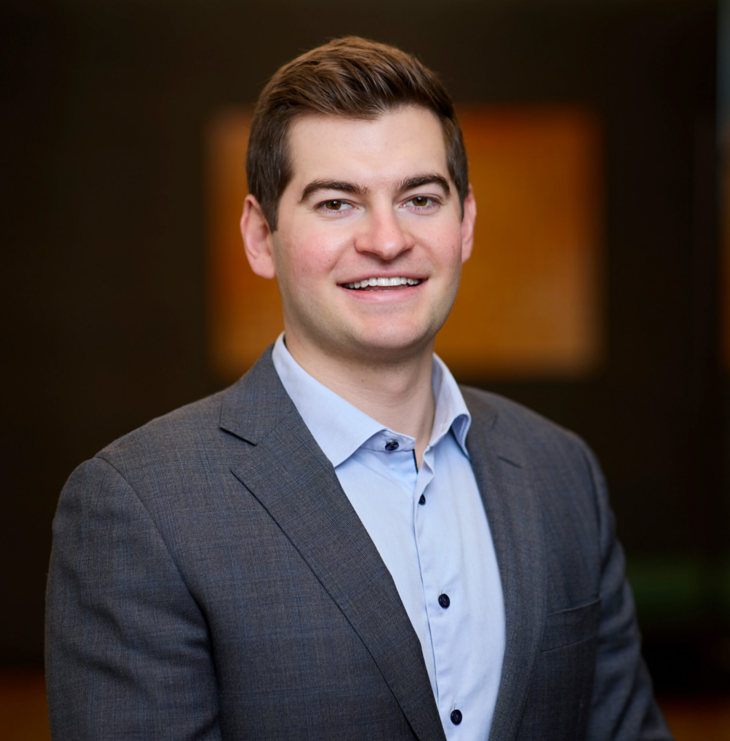 A man in a gray suit jacket and light blue shirt smiles at the camera against a blurred indoor background.