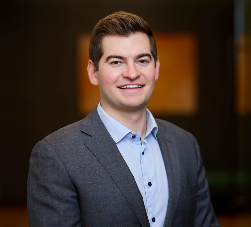 A man in a gray suit jacket and light blue shirt smiles at the camera against a blurred indoor background.