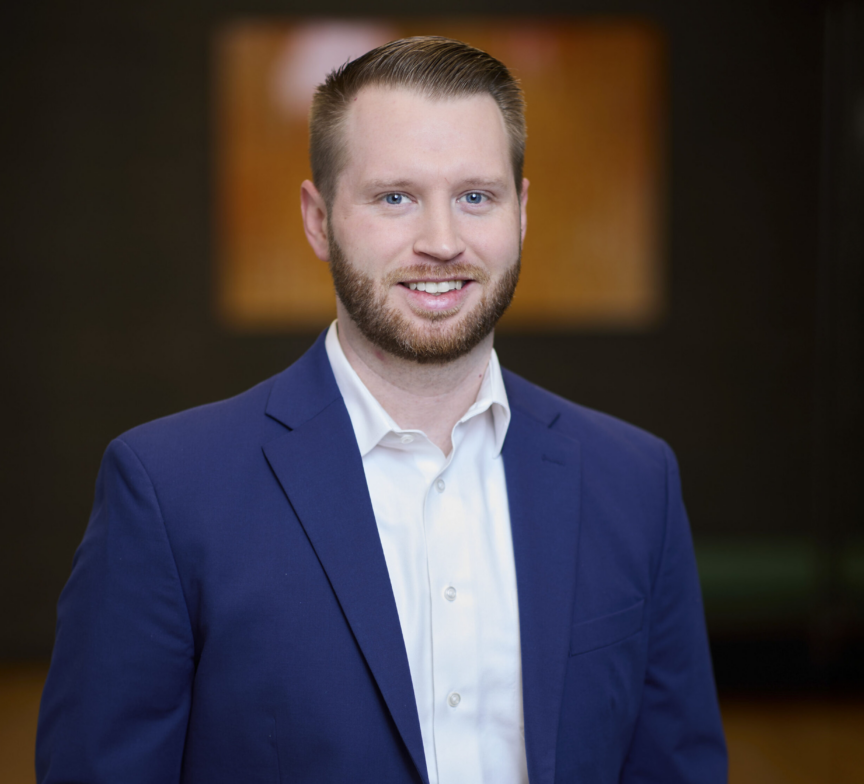 Andrew Brochu, a man with short light brown hair and a beard, is wearing a blue suit jacket and white dress shirt, smiling at the camera. The background is blurred and dark with a warm-colored rectangle behind him.