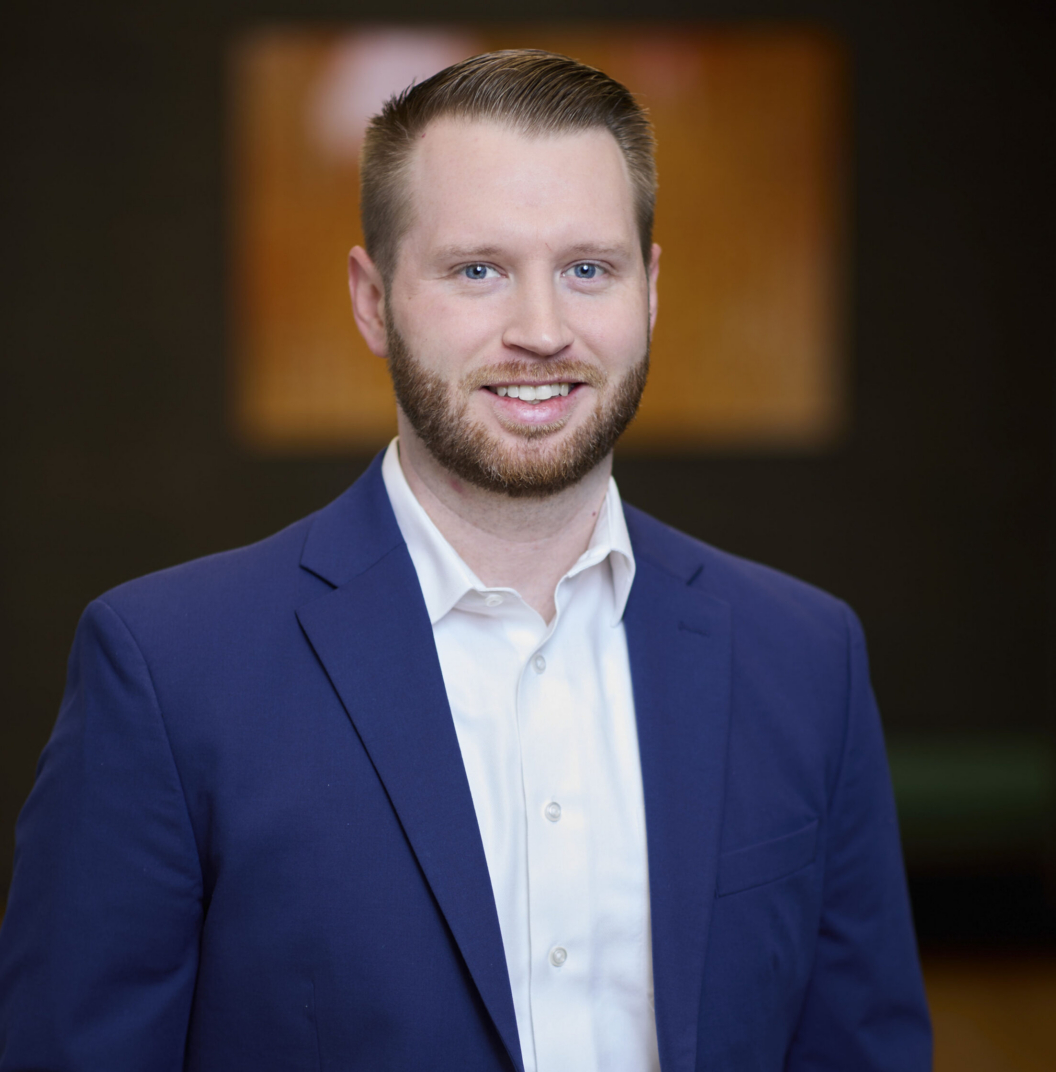 Andrew Brochu, a man with short light brown hair and a beard, is wearing a blue suit jacket and white dress shirt, smiling at the camera. The background is blurred and dark with a warm-colored rectangle behind him.