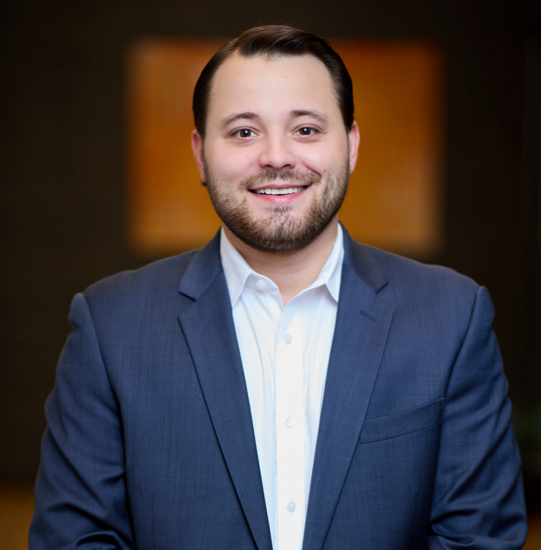 A man in a blue suit and white shirt smiles at the camera, standing indoors against a blurred dark background with warm light tones.