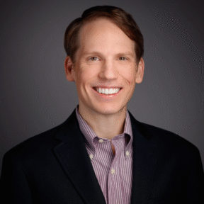 A smiling man with light brown hair wearing a black blazer over a striped button-down shirt, posing in front of a plain dark background.