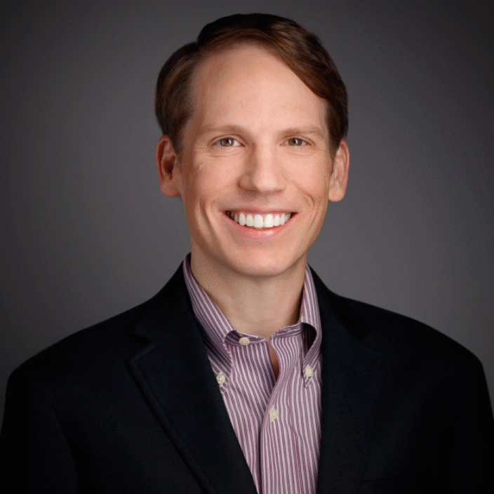 A smiling man with light brown hair wearing a black blazer over a striped button-down shirt, posing in front of a plain dark background.