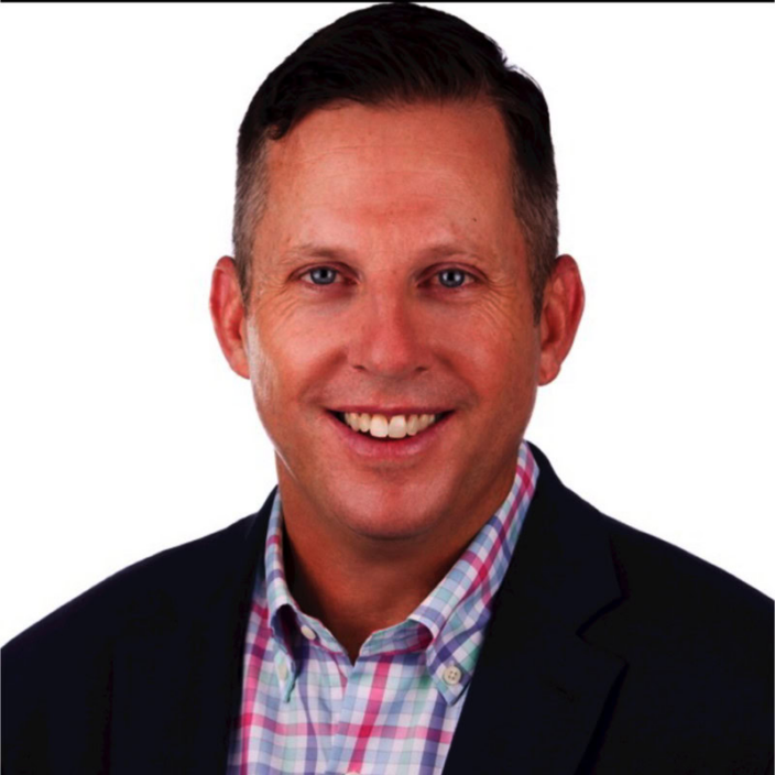 A man with short brown hair, wearing a dark blazer over a pink and blue plaid shirt, is smiling against a plain white background, representing Piper Sandler.