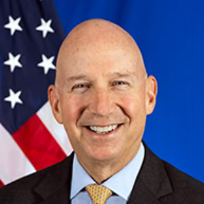 A bald man in a dark suit, light blue shirt, and yellow patterned tie smiles in front of a partial American flag and blue backdrop, embodying the confident demeanor of the Governor of Delaware.