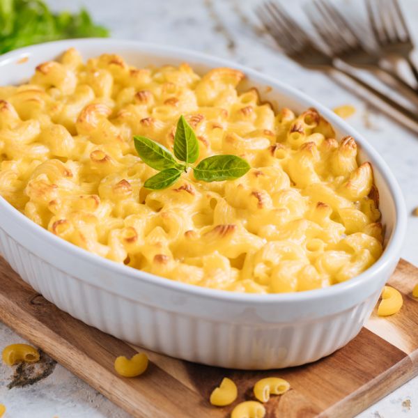 Baked macaroni and cheese in a white casserole dish, crafted with Revela Foods ingredients, is topped with a sprig of fresh basil and sits on a wooden board with forks and uncooked pasta scattered nearby.