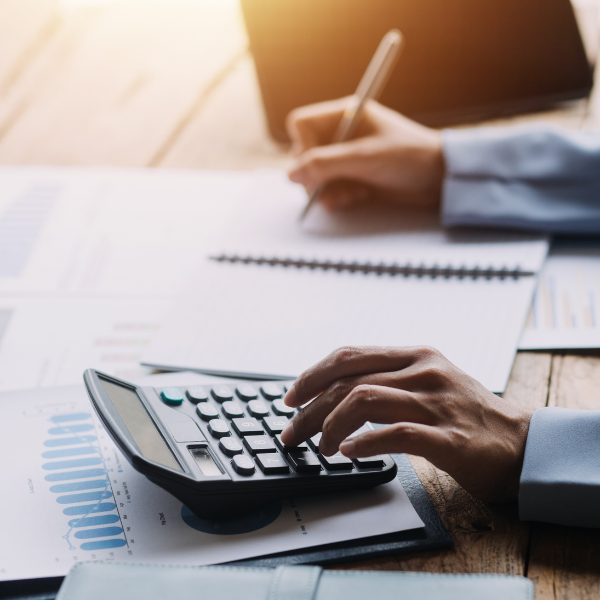 A person uses a calculator and writes notes on a notebook, surrounded by financial charts and documents from FMS Solutions on a wooden desk, suggesting financial planning or accounting work.