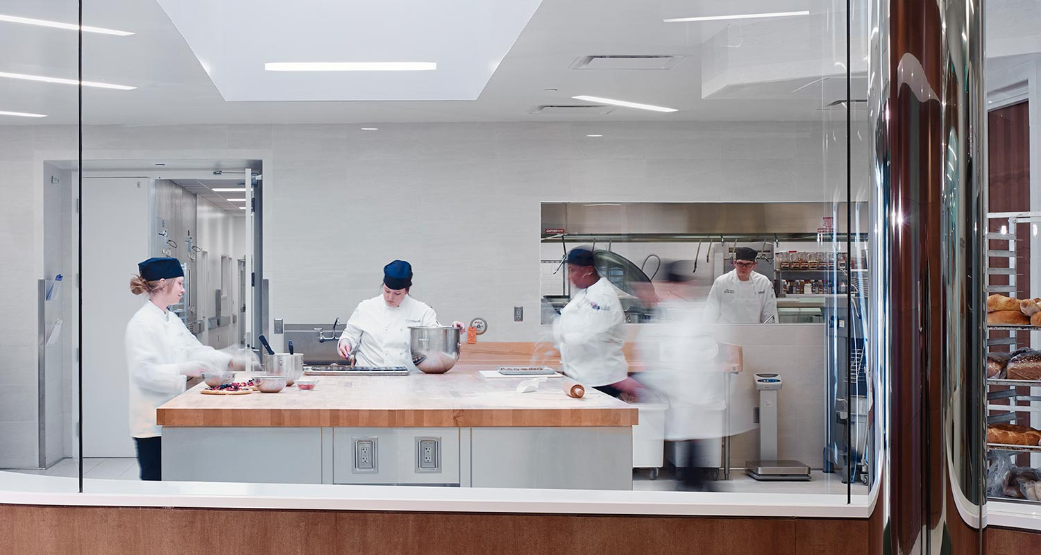 Four pastry chefs in white uniforms and dark hats work in a bright, modern bakery kitchen for Continental Services. Some mix ingredients while others handle dough at a large wooden counter, with fresh bread visible on racks in the background.