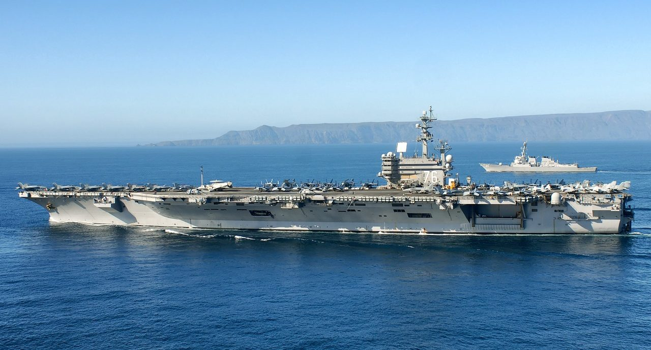 A large aircraft carrier sails on blue ocean water with numerous jets on its deck, accompanied by a smaller naval ship in the background, and an island is visible on the horizon under a clear sky.