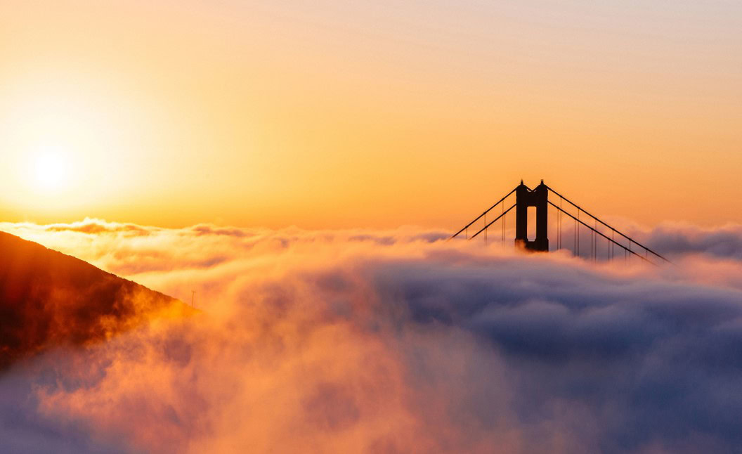 The sun rises over a thick layer of fog, partially obscuring the Golden Gate Bridge, with only the bridge towers and cables visible above the clouds.