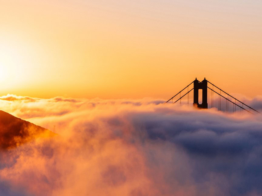 The sun rises over a thick layer of fog, partially obscuring the Golden Gate Bridge, with only the bridge towers and cables visible above the clouds.