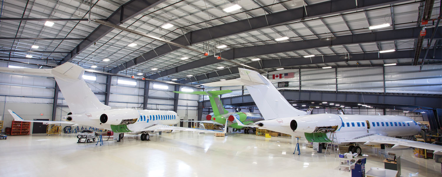 Two white private jets and one green jet, maintained by Flying Colours Corp, are parked inside a spacious, well-lit aircraft hangar with high ceilings, large windows, and visible maintenance equipment around the planes.