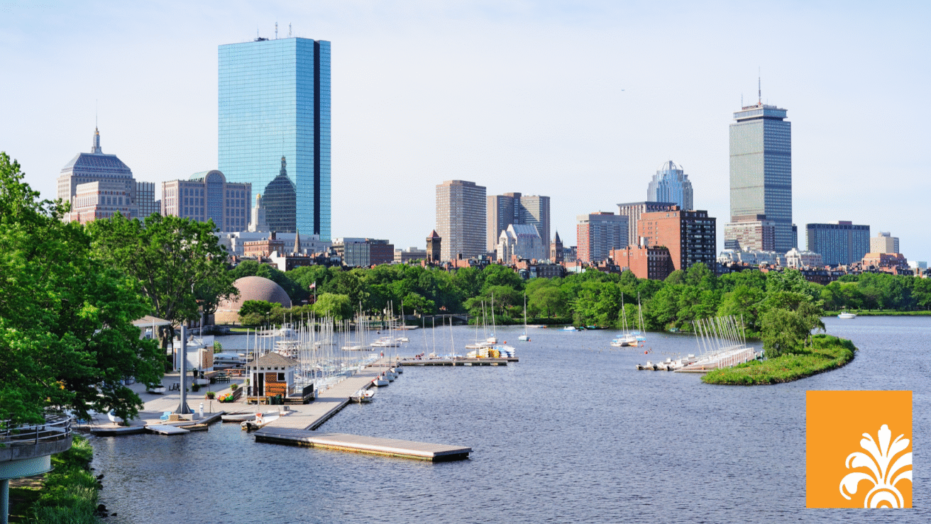A view of sailboats docked along the Charles River with the Boston skyline, including the Prudential and John Hancock Towers—home to firms like New Heritage Capital—visible in the background on a clear day.