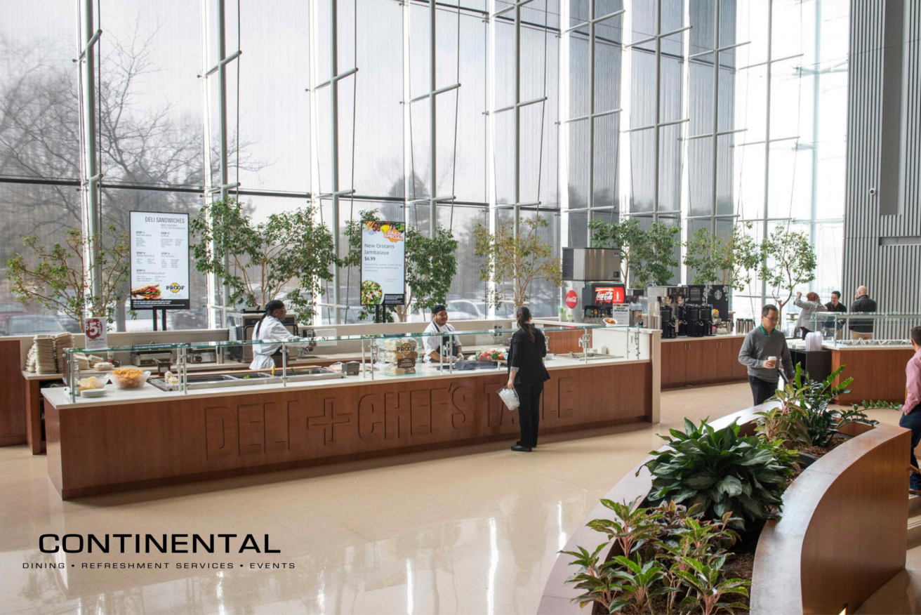 A modern cafeteria with large windows, light floors, and food counters staffed by employees. Several people are choosing food. Green plants and digital menu boards decorate the space. The Continental Services logo is visible in the corner.