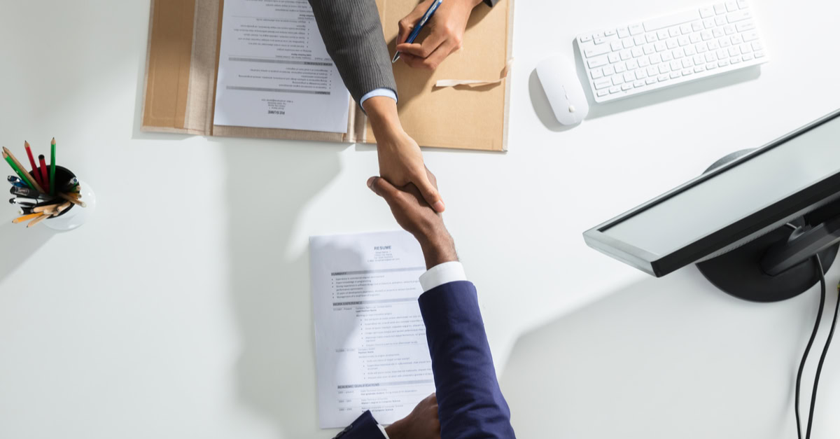 Two people are seated at a desk, shaking hands over documents, one labeled "resume." A computer monitor, keyboard, mouse, and a container of colored pencils sit on the white desk—typical of top offices in the best recruiting firms.