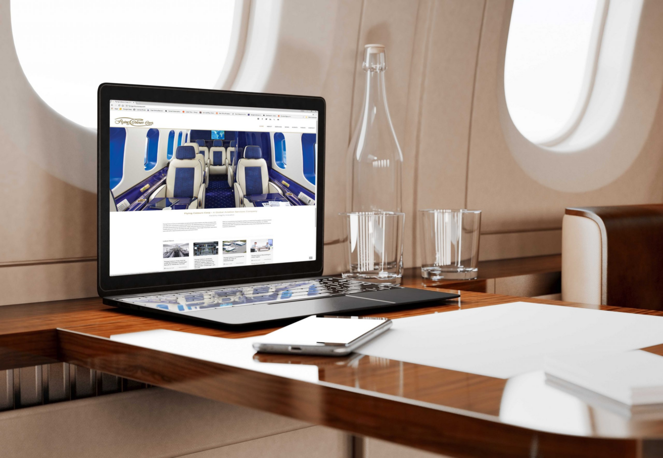 A laptop on a wooden table inside a private jet, showcasing high tech connectivity. Nearby are a glass water bottle, two glasses, a smartphone, and sheets of paper. Large windows provide natural light—an experience perfected by Flying Colours Corp.