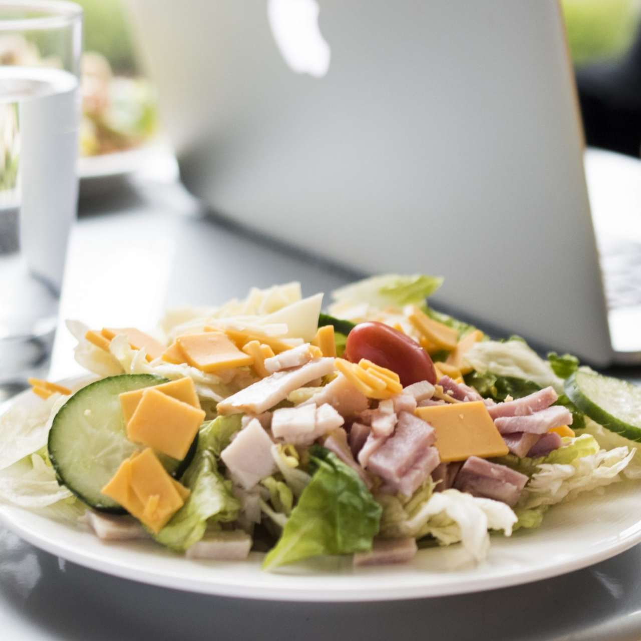 A plate of salad with lettuce, cucumber, tomato, cheese, and bacon sits next to a glass of water and an open laptop in a bright dining setting—ideal for busy employers or Continental Services’ meal breaks.